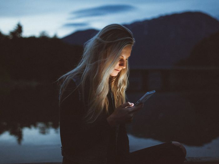 A woman sitting outdoors at dusk, her face lit by the phone she is reading